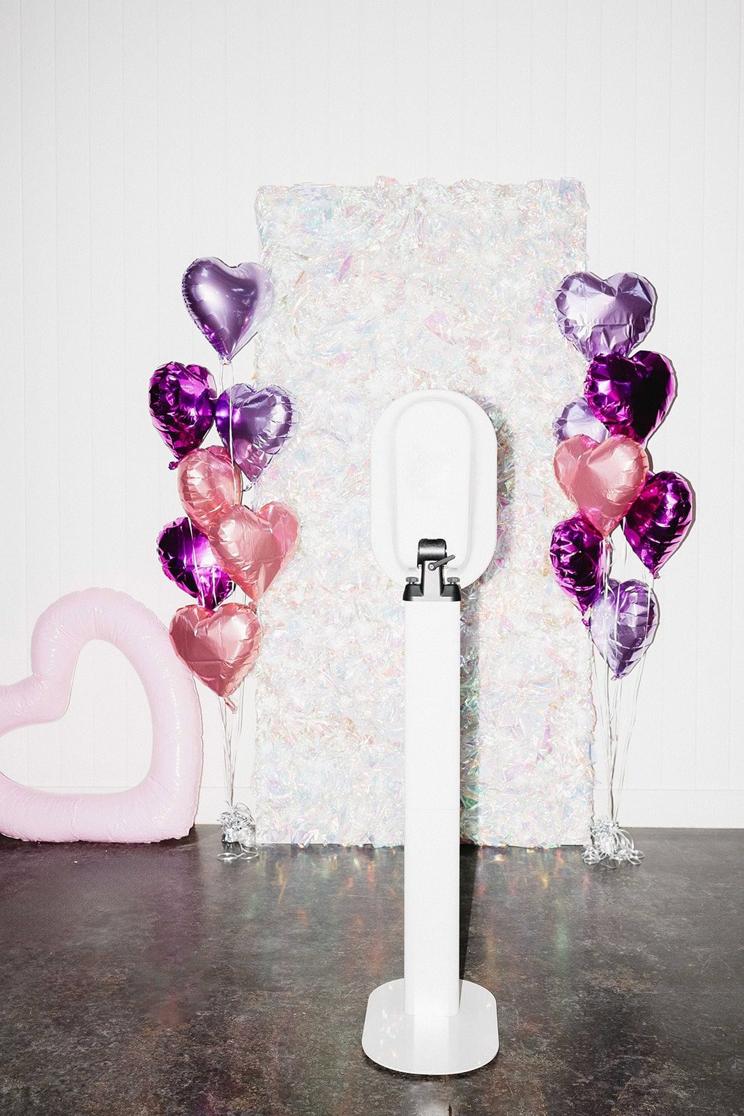 White photo booth kiosk before an iridescent backdrop flanked by pink and purple heart balloons