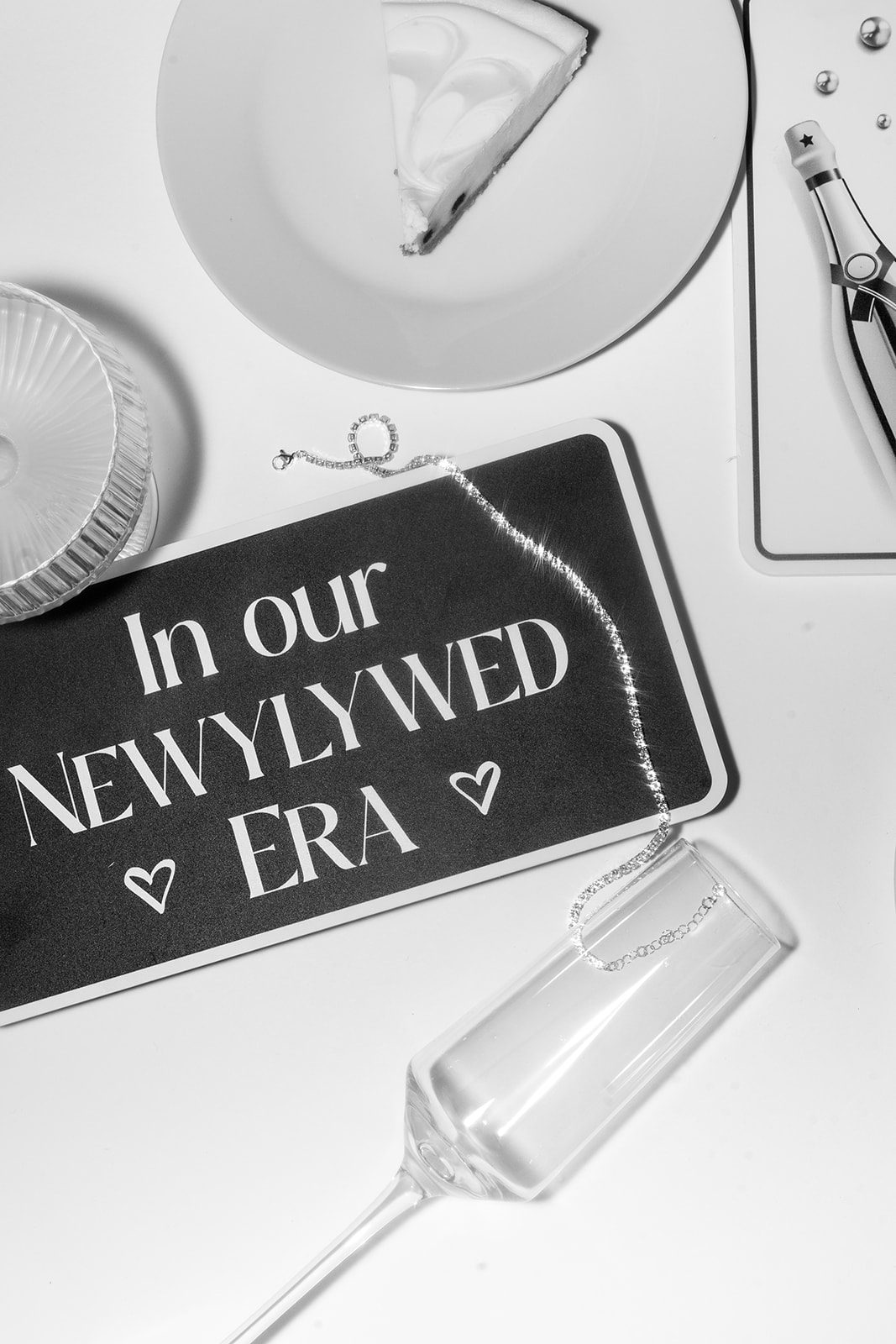 A black and white flat lay of an In Our Newlywed Era sign, a rhinestone chain and an empty champagne flute