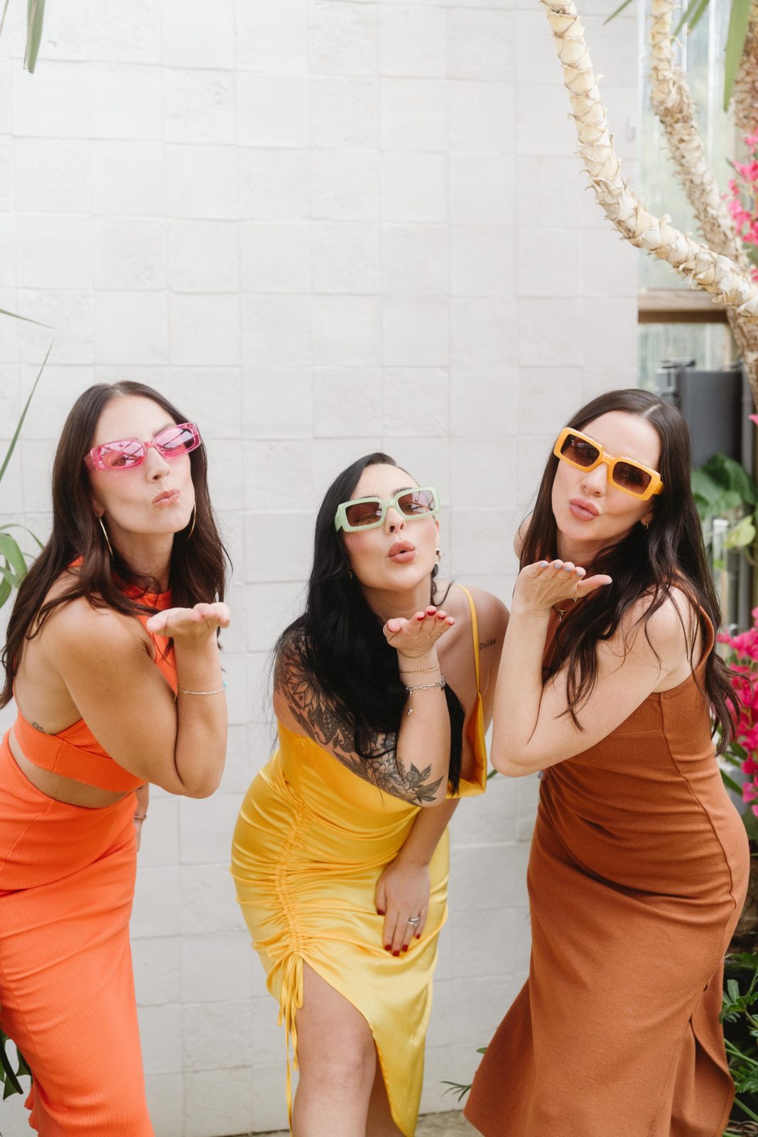 Three guests in orange, yellow and brown dresses wear colorful sunglasses and blow kisses