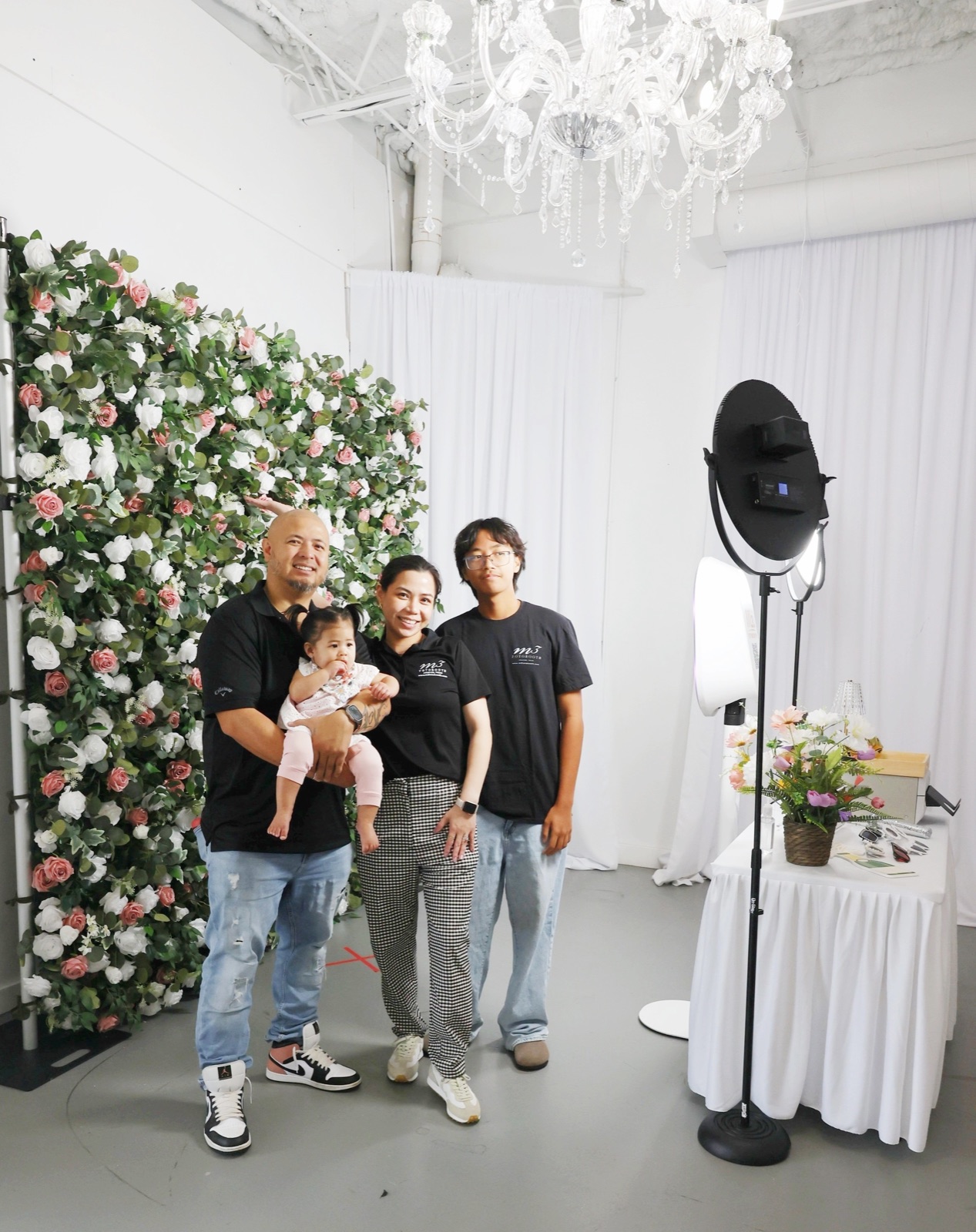 A man holding a baby, a woman and a young man by a rose flower wall and booth light
