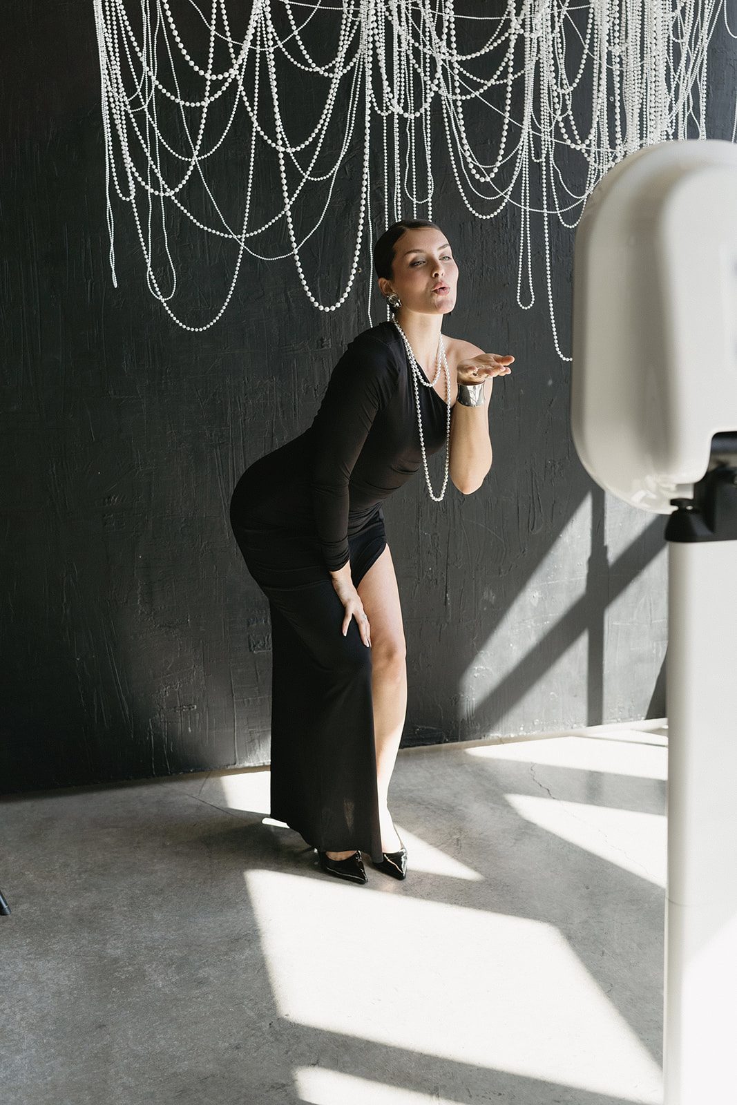 A guest in a long black gown blowing a kiss toward the booth, standing in a shaft of afternoon light beneath a beaded chandelier