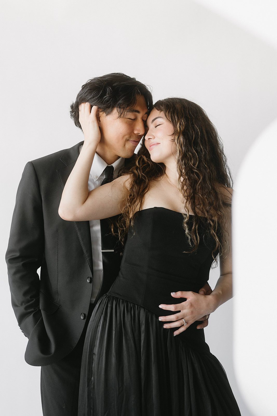 A couple in a black suit and black strapless gown stand with foreheads touching before a white wall